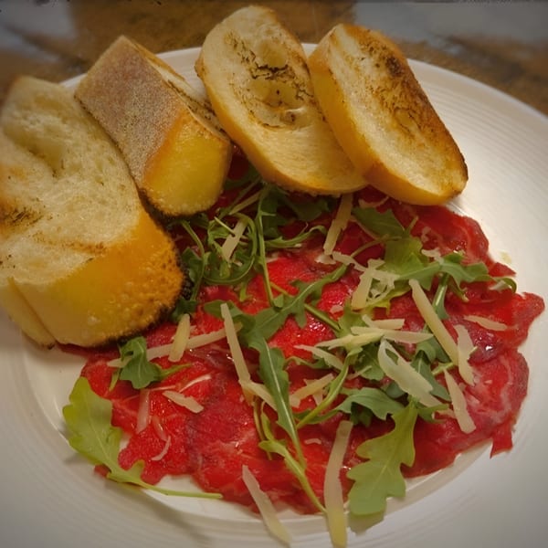 Beef carpaccio with arugula, shaved Parmesan, and toasted baguette slices on a white plate.