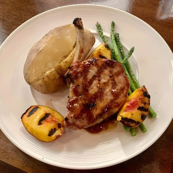 A glazed pork chop with a baked potato, grilled peaches, and asparagus on a white plate.